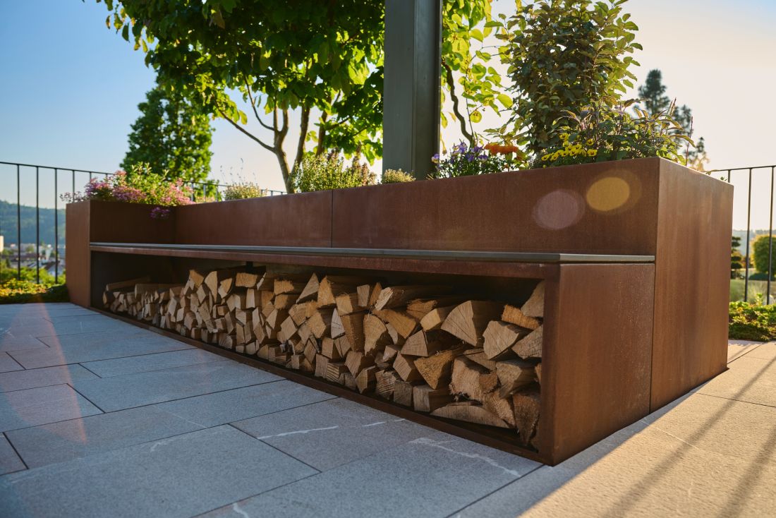 Modernes Hochbeet kombiniert mit Sitzbank und Lagerplatz für Feuerholz, bepflanzt mit blühenden Blumen und einem kleinen Baum