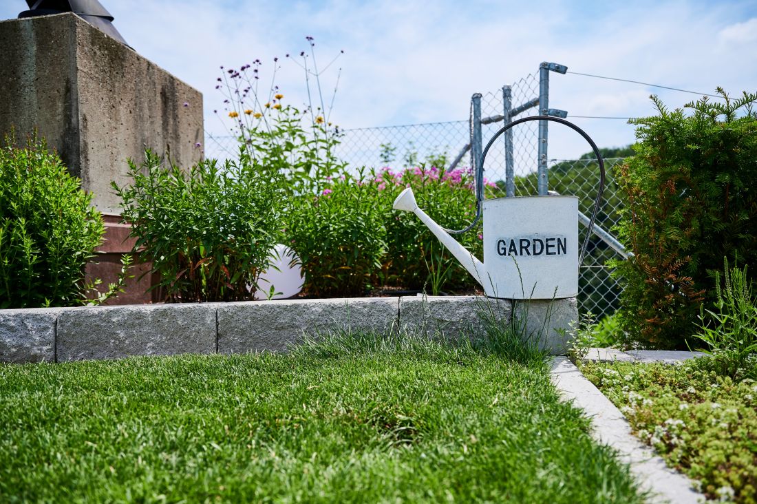 Weisser Giesskanne mit Schrift Garten vor gepflegtem Rasen und Blumenbeet Gartenbau Aargau Schoop Gartenbau