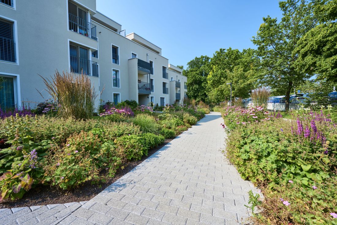 Gepflegte Gartenanlage einer Wohnüberbauung, links und rechts vom Weg bepflanzte Bette mit Stauden und verschiedenen Pflanzen