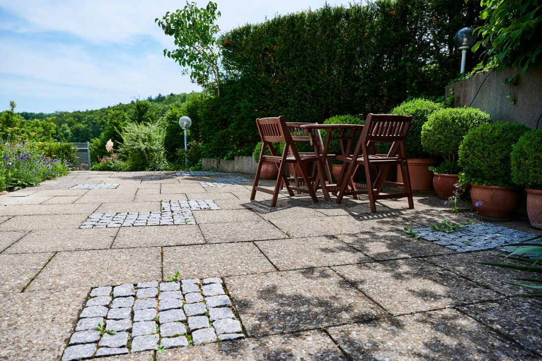 Terrassenbelag mit alten Gartenplatten, aufgelockert mit einzelnen Elementen aus Pflästersteinen, im Hintergrund ein Tische und Stühle aus Holz vor Pflanzkübeln und einer grünen Hecke