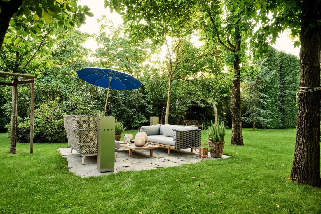 Stylischer Gartensitzplatz in Privatgarten, umgeben mit Bäumen, im Morgenlicht, Gartenlounge und -tisch auf Steinplatten