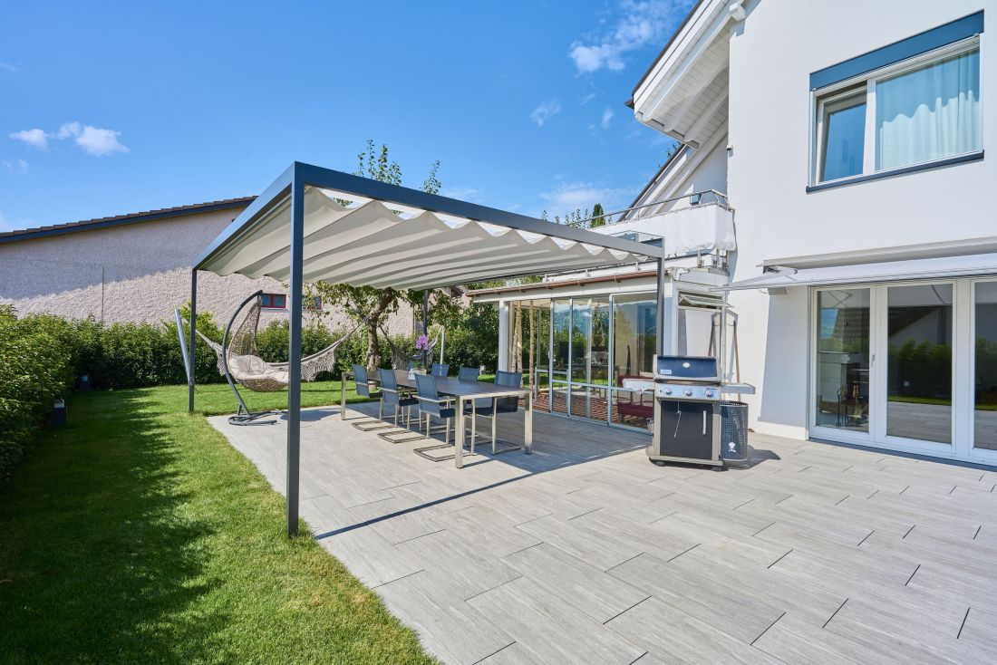 Moderner gedeckter Sitzplatz in einem Privatgarten im Aargau, Esstisch mit Stühlen, Grill und gemütlichem Hängesessel