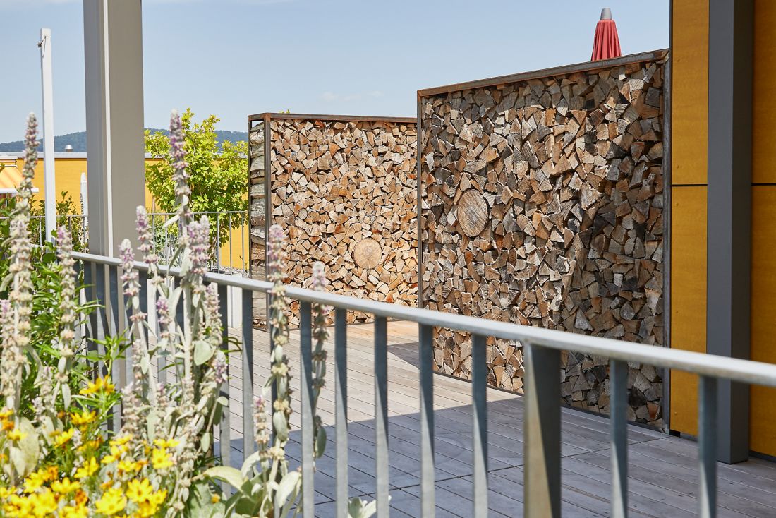 Terrassengestaltung mit Holzstapel als Sichtschutz und blühenden Pflanzen, Gartenbau Aargau Schoop Gartenbau