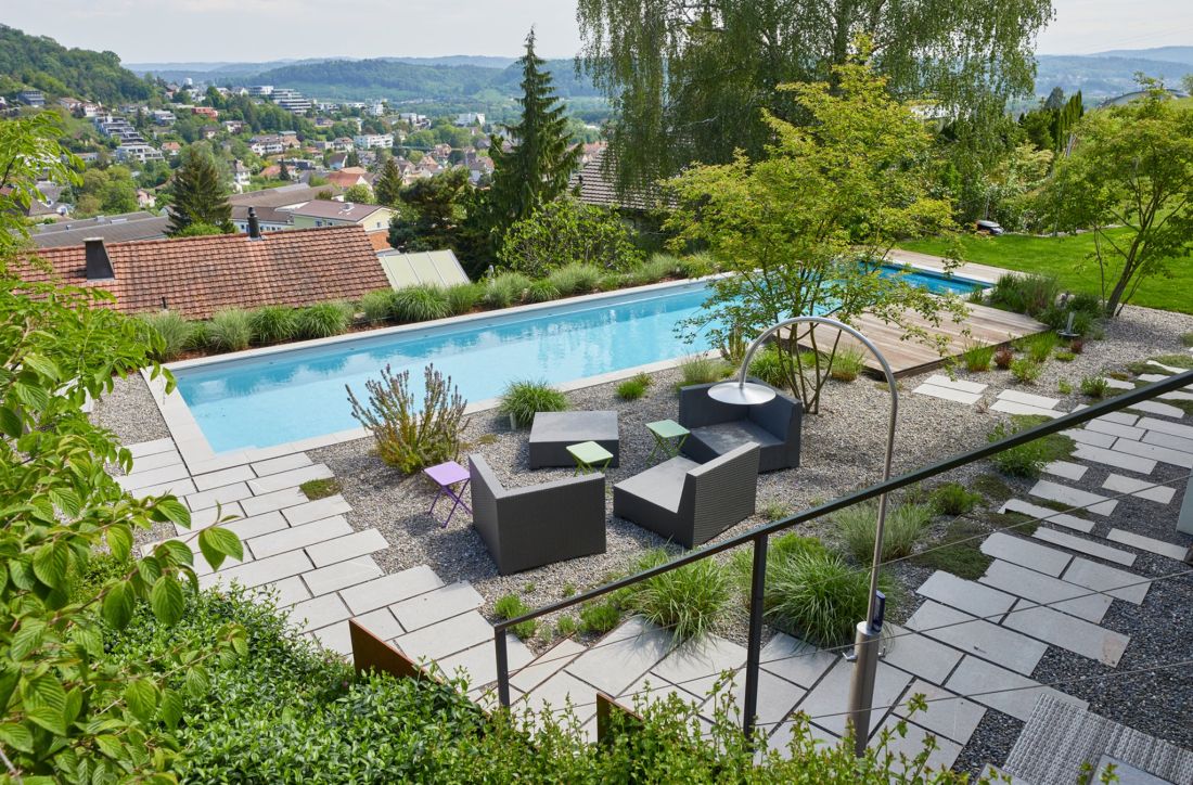 Grosser Garten mi Pool, umgeben von Steinplatten und Kiesplatz mit Sitzlounge und Liegefläche aus Holz