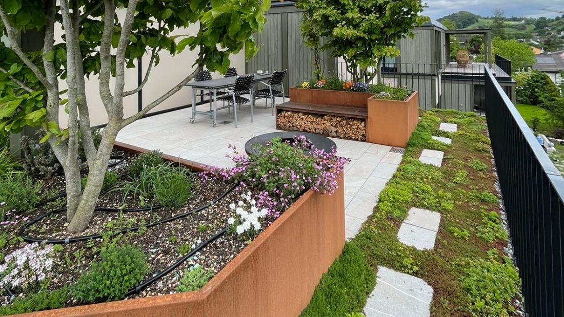 Terrasse mit bepflanzten Hochbeeten und automatischer Bewässerung (schwarzer Schlauch), Terrasse mit Gartenplatten und Esstisch