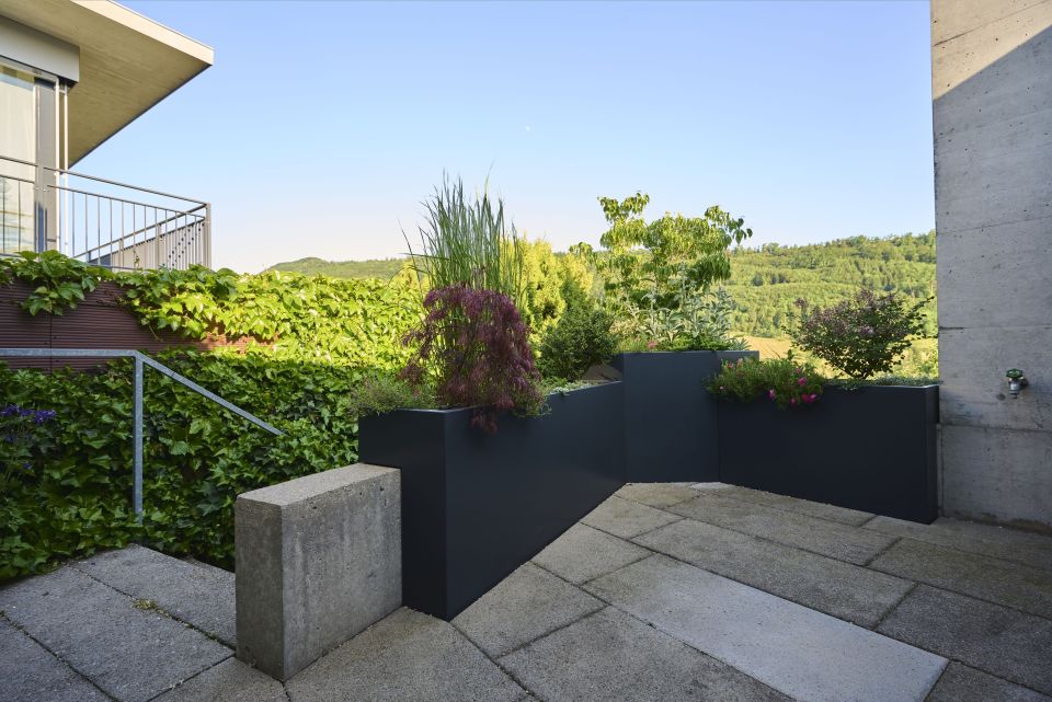 Terrassenabgrenzung mit bepflanzten Trögen, Steinplatten, Treppengeländer und Hecke im Hintergrund