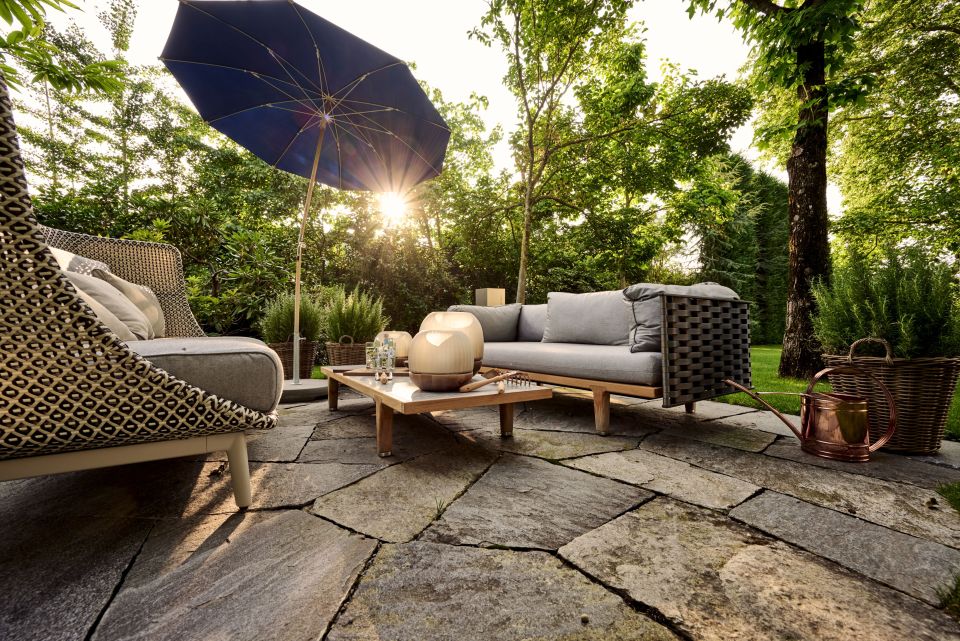 Moderne Gartensitzgruppe mit Polstersofa, blauem Sonnenschirm und Pflanzen auf Natursteinplatten, stimmige Abendsonne