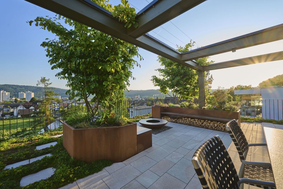 Moderne Terrasse mit aus rostigem Stahl eingefassten Hochbeeten, die mit kleinen Bäumen und verschiedenen Blumen bepflanzt sind, in der Mitte ist eine runde Grillstelle
