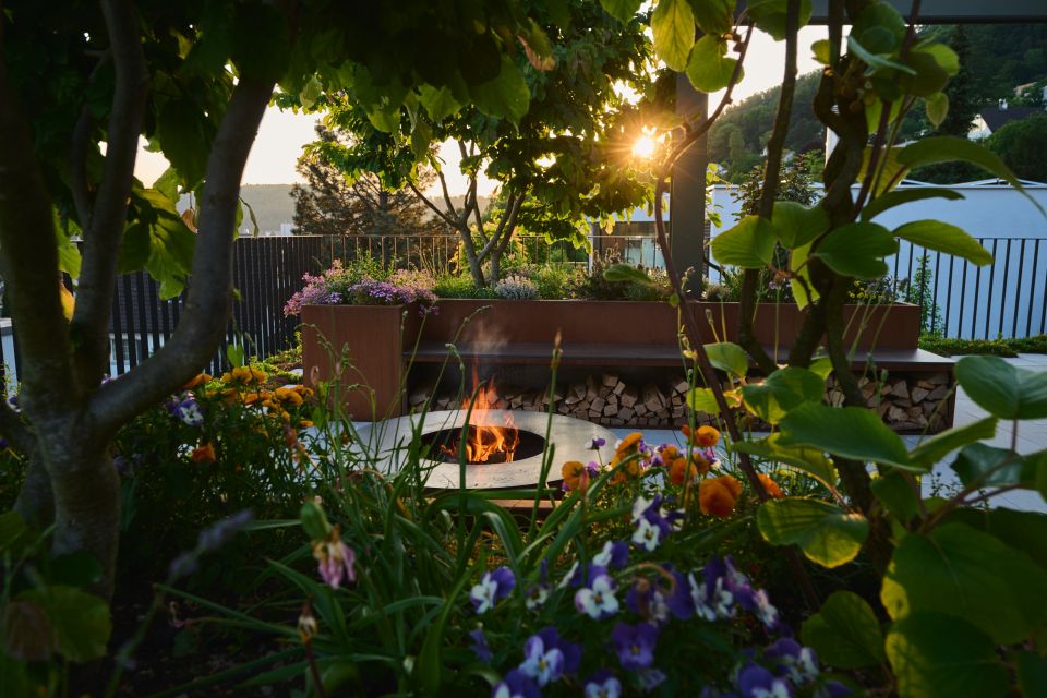 Runde Grillschale mit Flamme in der Abendsonne, Blick durch bunte Pflanzen, im Hintergrund ein bepflanztes Hochbett und die Abendsonne, die durchscheint