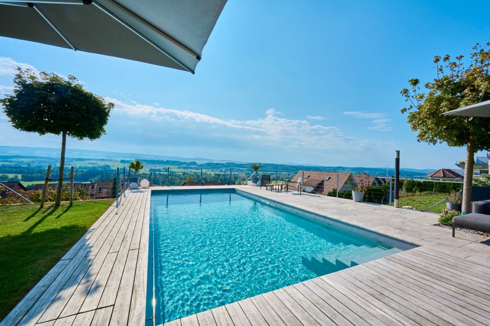Swimmingpool umrandet von einem hellen Holzdeck, links einen kleine Rasenfläche mit Bäumchen, im Hintergrund blauer Himmel