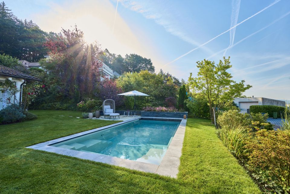 Pool in einem Privatgarten, umrandet von Granitplatten und gepflegtem grünen Rasen. Angrenzend ein Sitzplatz mit stilvollem Liegemöbel und Sonnenschirm