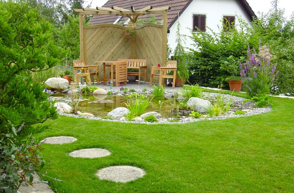 Gartenteich mit Wasserpflanzen, Steinen und Kiesumrandung, im Hintergrund ein Sitzplatz mit Sichtschutz aus Holz, im Vordergrund eine gepflegte Rasenfläche mit runden Steinplatten als Gehweg