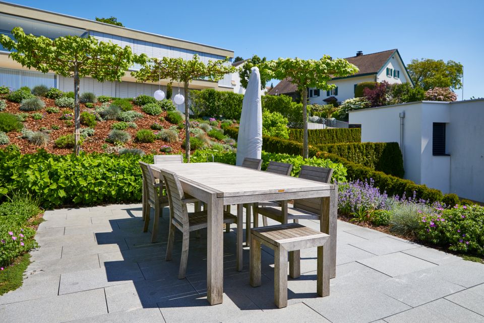 Moderner Gartensitzplatz mit Holztisch und Stühlen, Bodenbelag aus Steinplatten, umgeben von bepflanzten Beeten mit verschiedenen Grüntönen und kleinen Bäumen
